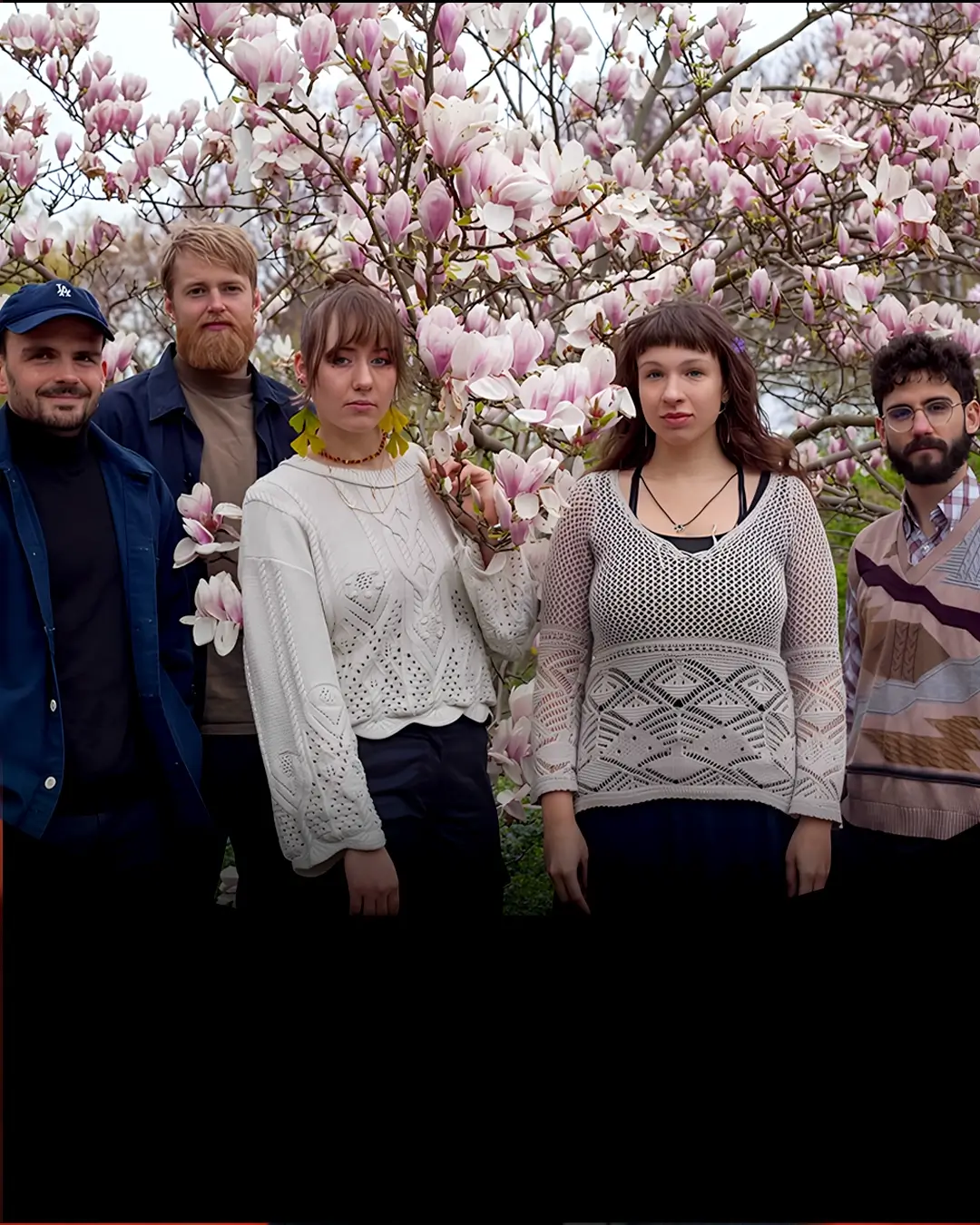 Five musicians stand together in front of a blooming magnolia tree, surrounded by pink flowers, looking calmly at the camera.