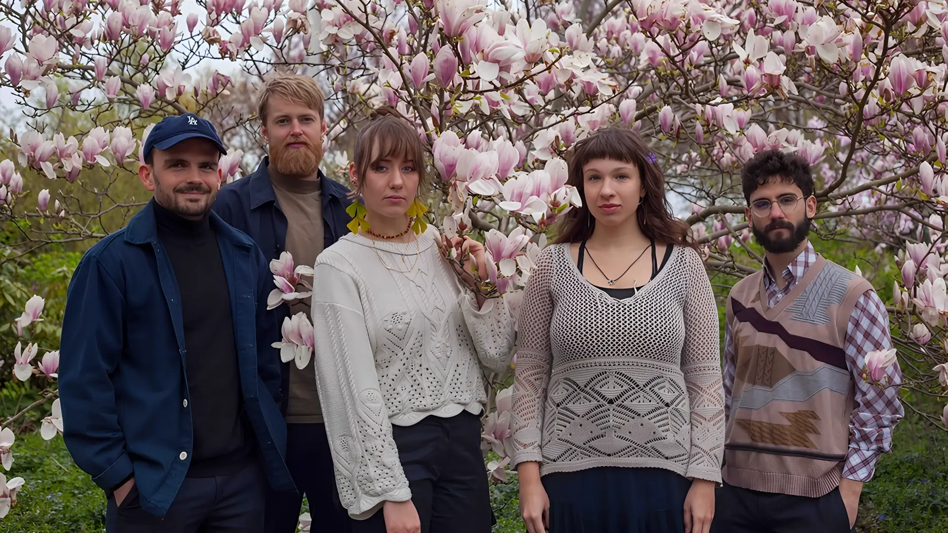 Five musicians stand together in front of a blooming magnolia tree, surrounded by pink flowers, looking calmly at the camera.