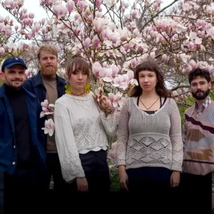 Five musicians stand together in front of a blooming magnolia tree, surrounded by pink flowers, looking calmly at the camera.