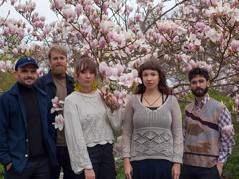Five musicians stand together in front of a blooming magnolia tree, surrounded by pink flowers, looking calmly at the camera.