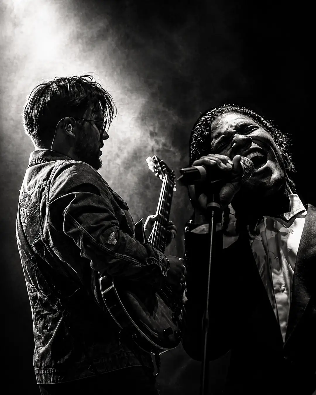 Zwart-wit podiumfoto van gitarist Lorijn van Piekartz die elektrische gitaar speelt naast zanger Jared Grant, die krachtig in de microfoon zingt, omgeven door dramatisch licht en rook.