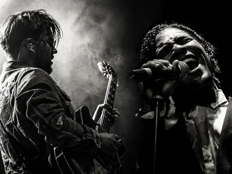 Zwart-wit podiumfoto van gitarist Lorijn van Piekartz die elektrische gitaar speelt naast zanger Jared Grant, die krachtig in de microfoon zingt, omgeven door dramatisch licht en rook.