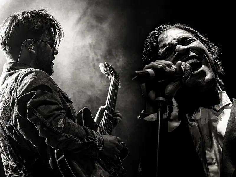 Zwart-wit podiumfoto van gitarist Lorijn van Piekartz die elektrische gitaar speelt naast zanger Jared Grant, die krachtig in de microfoon zingt, omgeven door dramatisch licht en rook.