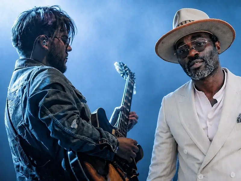Optredenscène met gitarist Lorijn van Piekartz en zanger Steffen Morrison op het podium, omringd door blauw licht en lichte rook; de gitarist speelt elektrisch terwijl de zanger in een licht pak en hoed richting publiek kijkt.