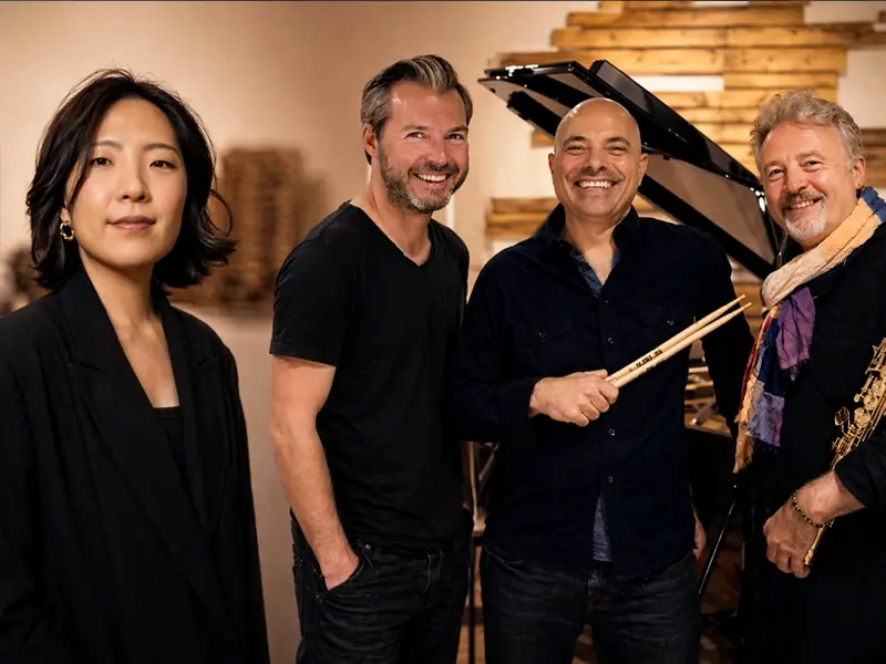 Miran Noh and members of the Tim Garland Lighthouse Trio posing together beside a grand piano in a studio, holding instruments and smiling.