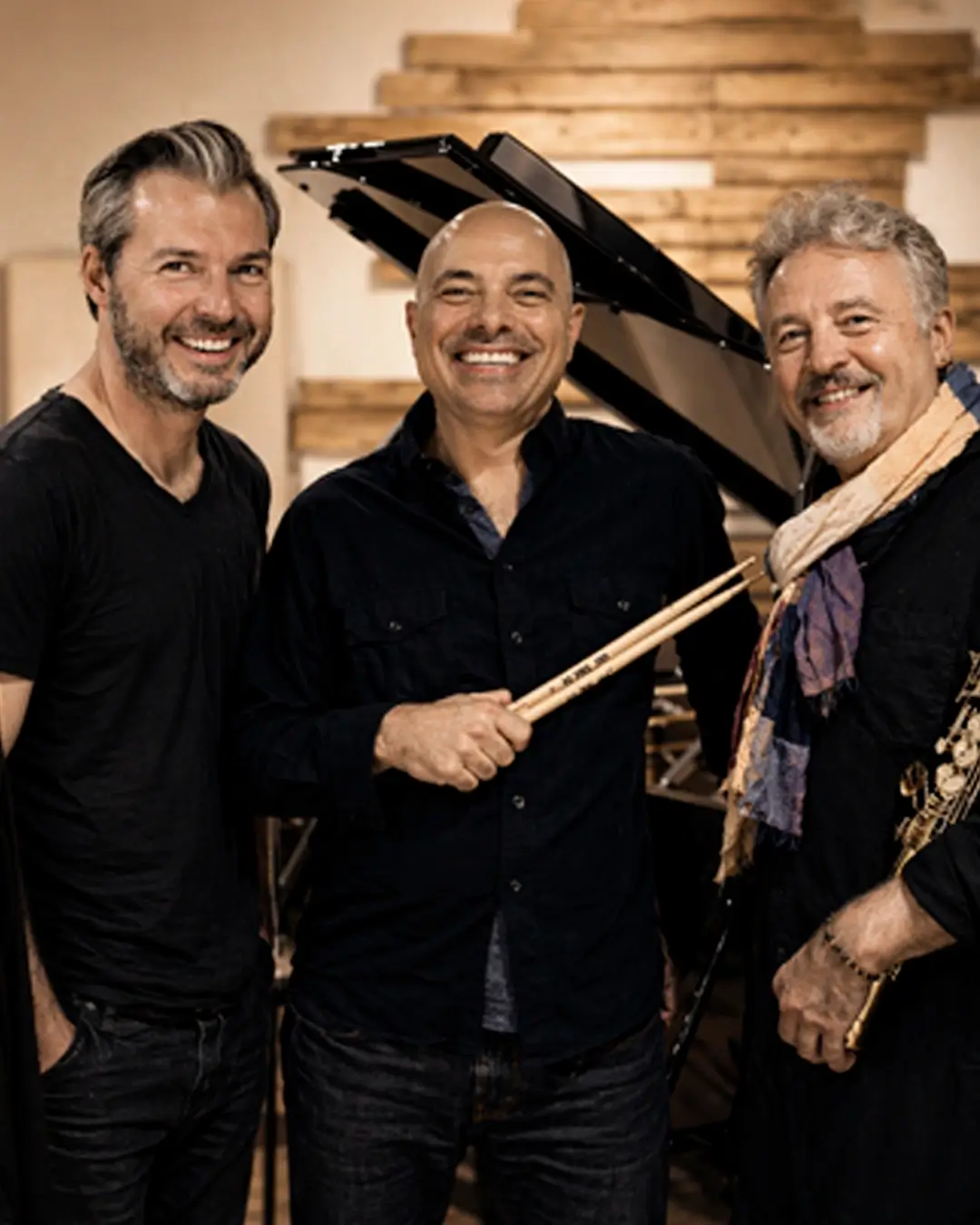 Drie jazzmuzikanten van het Tim Garland Lighthouse Trio – saxofonist, pianist en drummer – lachend bij een vleugel in een studio.