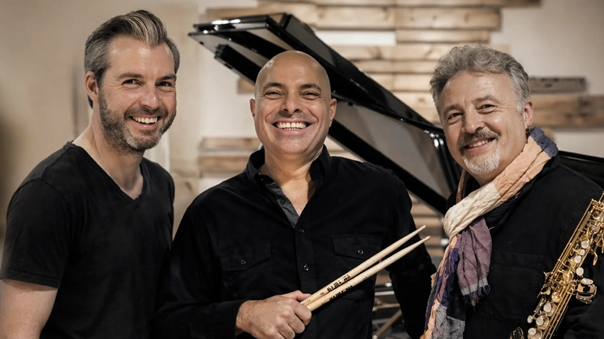 Drie jazzmuzikanten van het Tim Garland Lighthouse Trio – saxofonist, pianist en drummer – lachend bij een vleugel in een studio.