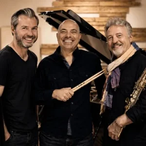 Drie jazzmuzikanten van het Tim Garland Lighthouse Trio – saxofonist, pianist en drummer – lachend bij een vleugel in een studio.