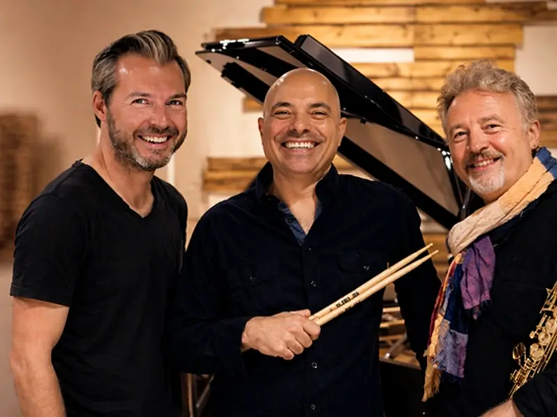 Drie jazzmuzikanten van het Tim Garland Lighthouse Trio – saxofonist, pianist en drummer – lachend bij een vleugel in een studio.