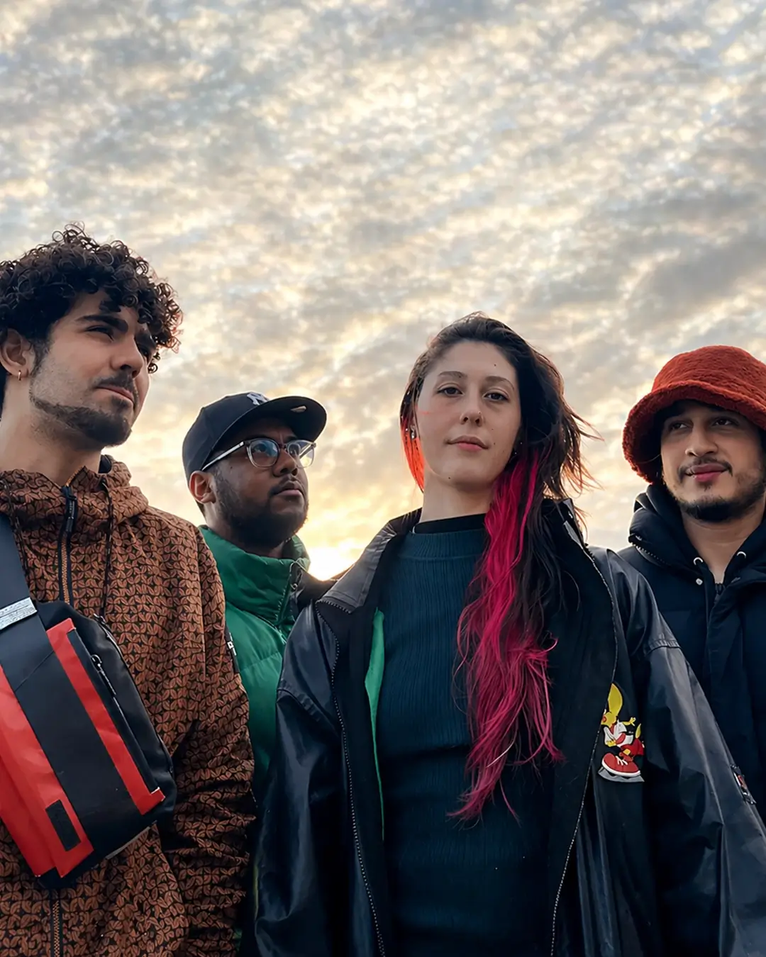 Four young musicians stand together in mixed shadow and sunlight, looking at the camera against a dark background with strong contrasts.