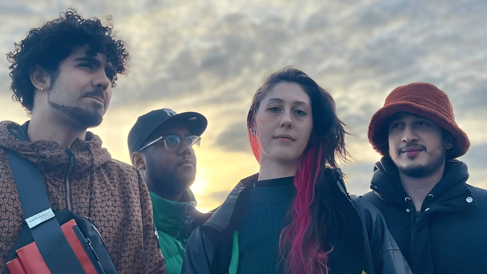 Four young musicians stand together in mixed shadow and sunlight, looking at the camera against a dark background with strong contrasts.