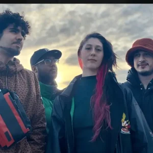 Four young musicians stand together in mixed shadow and sunlight, looking at the camera against a dark background with strong contrasts.