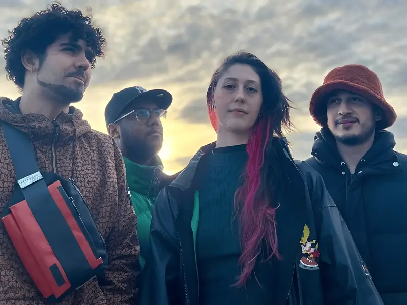 Four young musicians stand together in mixed shadow and sunlight, looking at the camera against a dark background with strong contrasts.