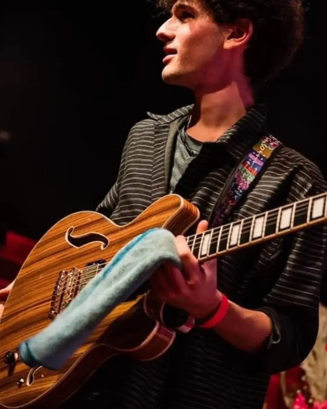 Agus Fulka op een podium met rood verlichte stoelen op de achtergrond, die een semi-hollow elektrische gitaar vasthoudt en zijwaarts kijkt terwijl hij zich voorbereidt om te spelen.