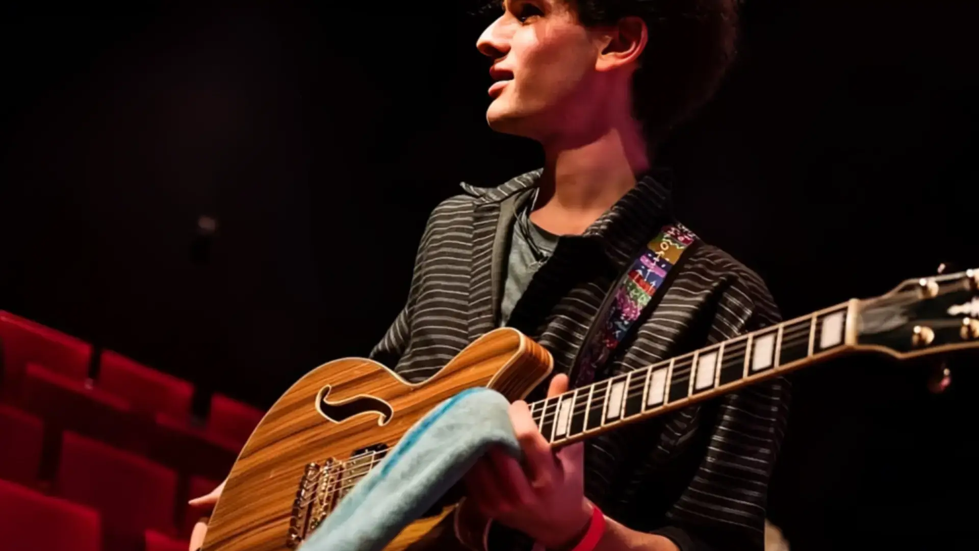 Agus Fulka op een podium met rood verlichte stoelen op de achtergrond, die een semi-hollow elektrische gitaar vasthoudt en zijwaarts kijkt terwijl hij zich voorbereidt om te spelen.