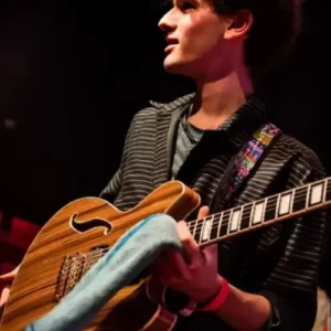 Agus Fulka op een podium met rood verlichte stoelen op de achtergrond, die een semi-hollow elektrische gitaar vasthoudt en zijwaarts kijkt terwijl hij zich voorbereidt om te spelen.