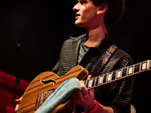 Agus Fulka op een podium met rood verlichte stoelen op de achtergrond, die een semi-hollow elektrische gitaar vasthoudt en zijwaarts kijkt terwijl hij zich voorbereidt om te spelen.