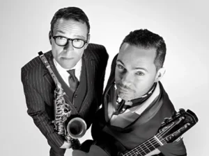 Zwart-wit portret van 
Benjamin Herman en Robin Nolan die omhoog in de camera kijken; links een saxofonist in pak met bril die zijn instrument vasthoudt, rechts een gitarist met een gitaar, beiden strak gekleed en close bij elkaar in beeld.