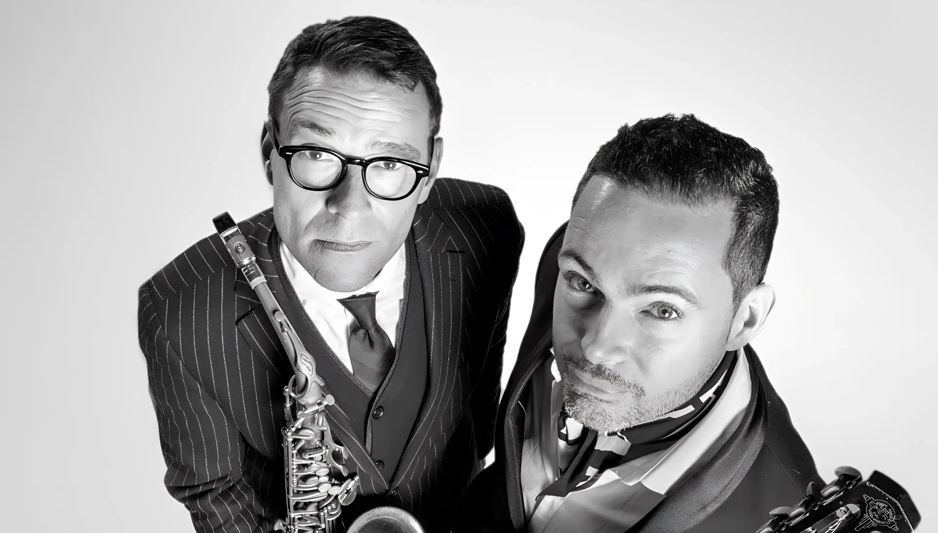 Zwart-wit portret van Benjamin Herman en Robin Nolan die omhoog in de camera kijken; links een saxofonist in pak met bril die zijn instrument vasthoudt, rechts een gitarist met een gitaar, beiden strak gekleed en close bij elkaar in beeld.