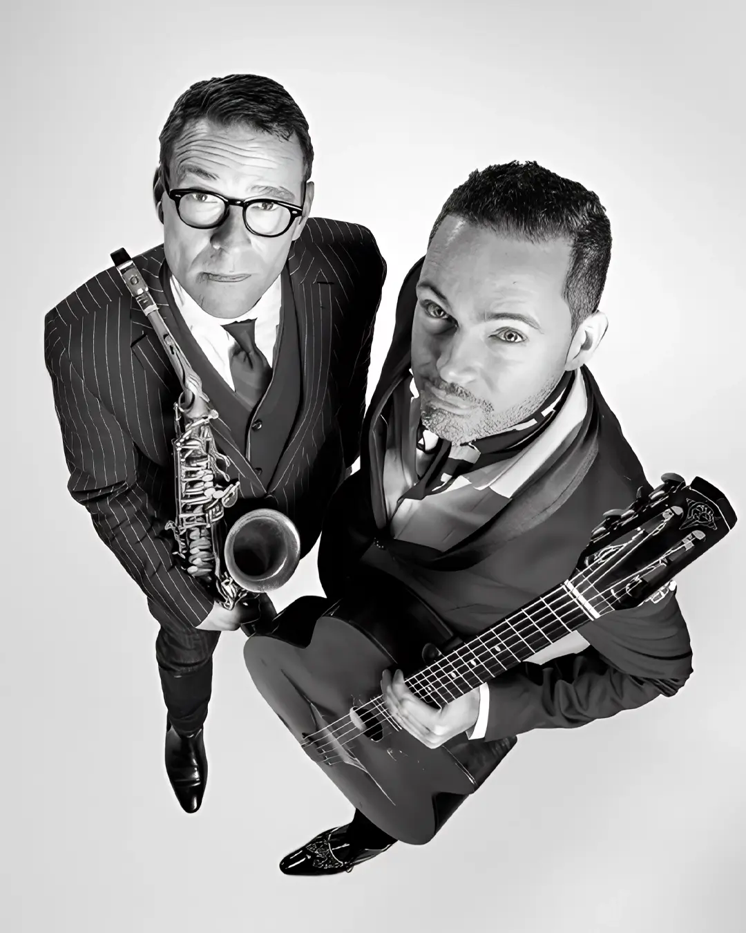 Zwart-wit portret van Benjamin Herman en Robin Nolan die omhoog in de camera kijken; links een saxofonist in pak met bril die zijn instrument vasthoudt, rechts een gitarist met een gitaar, beiden strak gekleed en close bij elkaar in beeld.
