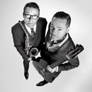 Zwart-wit portret van
Benjamin Herman en Robin Nolan die omhoog in de camera kijken; links een saxofonist in pak met bril die zijn instrument vasthoudt, rechts een gitarist met een gitaar, beiden strak gekleed en close bij elkaar in beeld.