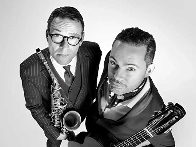 Zwart-wit portret van 
Benjamin Herman en Robin Nolan die omhoog in de camera kijken; links een saxofonist in pak met bril die zijn instrument vasthoudt, rechts een gitarist met een gitaar, beiden strak gekleed en close bij elkaar in beeld.