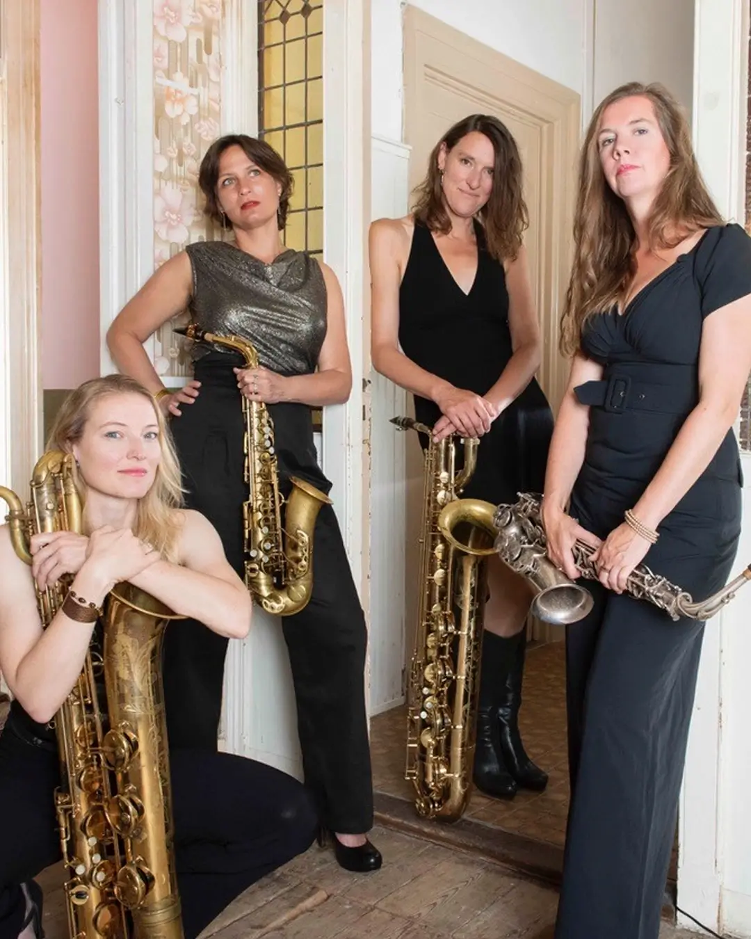 Vier vrouwelijke saxofonisten van het jazzkwartet Double Espresso poseren in een stijlvolle binnenruimte met hun instrumenten, waaronder alt- en baritonsaxofoons.