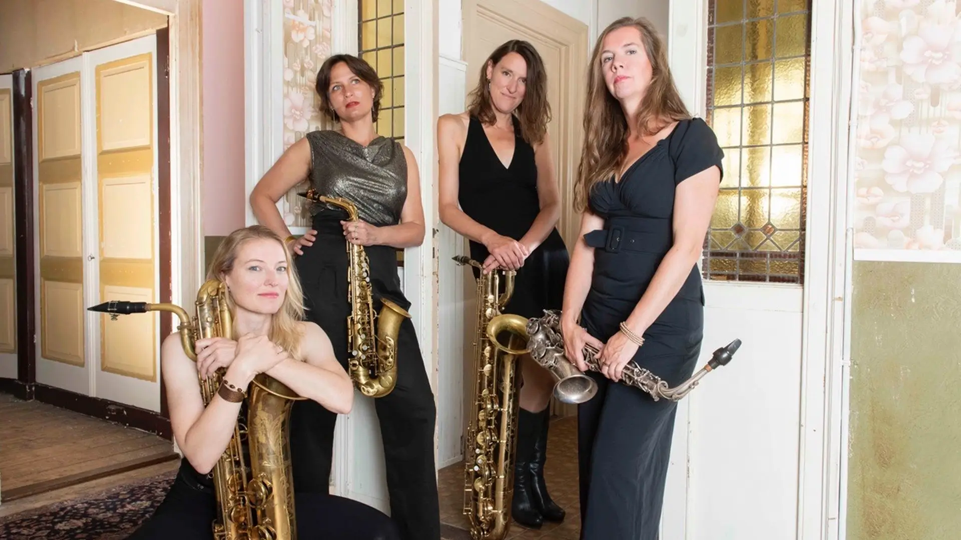 Vier vrouwelijke saxofonisten van het jazzkwartet Double Espresso poseren in een stijlvolle binnenruimte met hun instrumenten, waaronder alt- en baritonsaxofoons.