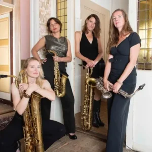 Vier vrouwelijke saxofonisten van het jazzkwartet Double Espresso poseren in een stijlvolle binnenruimte met hun instrumenten, waaronder alt- en baritonsaxofoons.