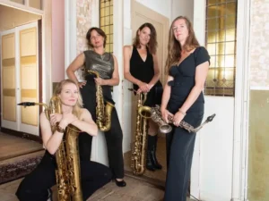 Vier vrouwelijke saxofonisten van het jazzkwartet Double Espresso poseren in een stijlvolle binnenruimte met hun instrumenten, waaronder alt- en baritonsaxofoons.