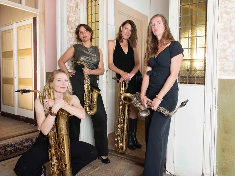 Vier vrouwelijke saxofonisten van het jazzkwartet Double Espresso poseren in een stijlvolle binnenruimte met hun instrumenten, waaronder alt- en baritonsaxofoons.