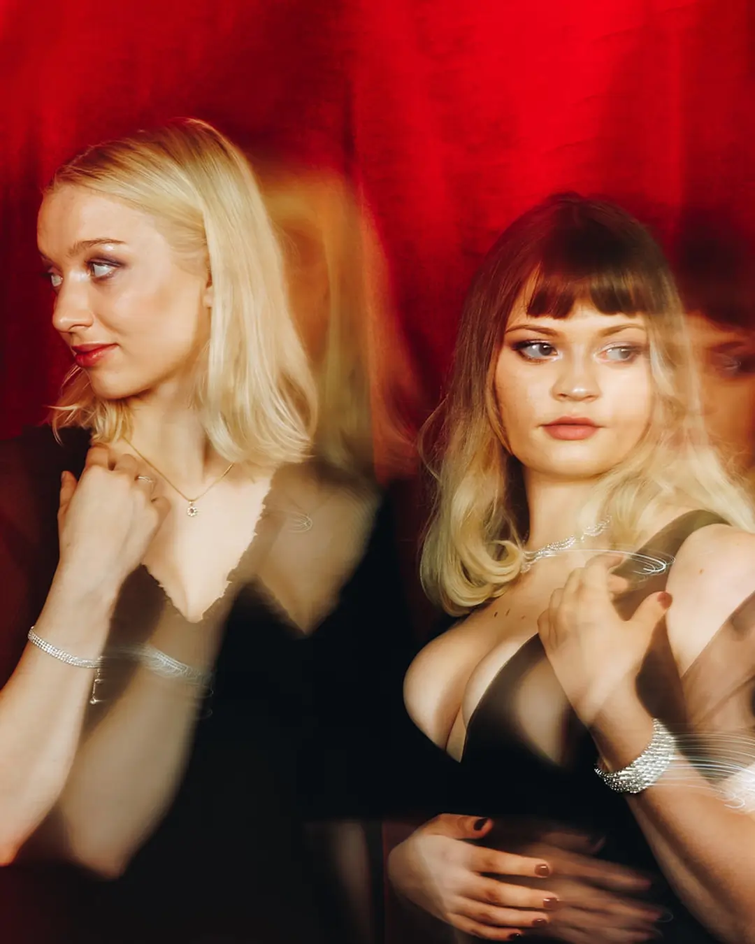 Twee zangeressen staan naast elkaar voor een rode achtergrond, stijlvol gekleed in zwart, met een dromerige en theatrale uitstraling door lichte bewegingsonscherpte.