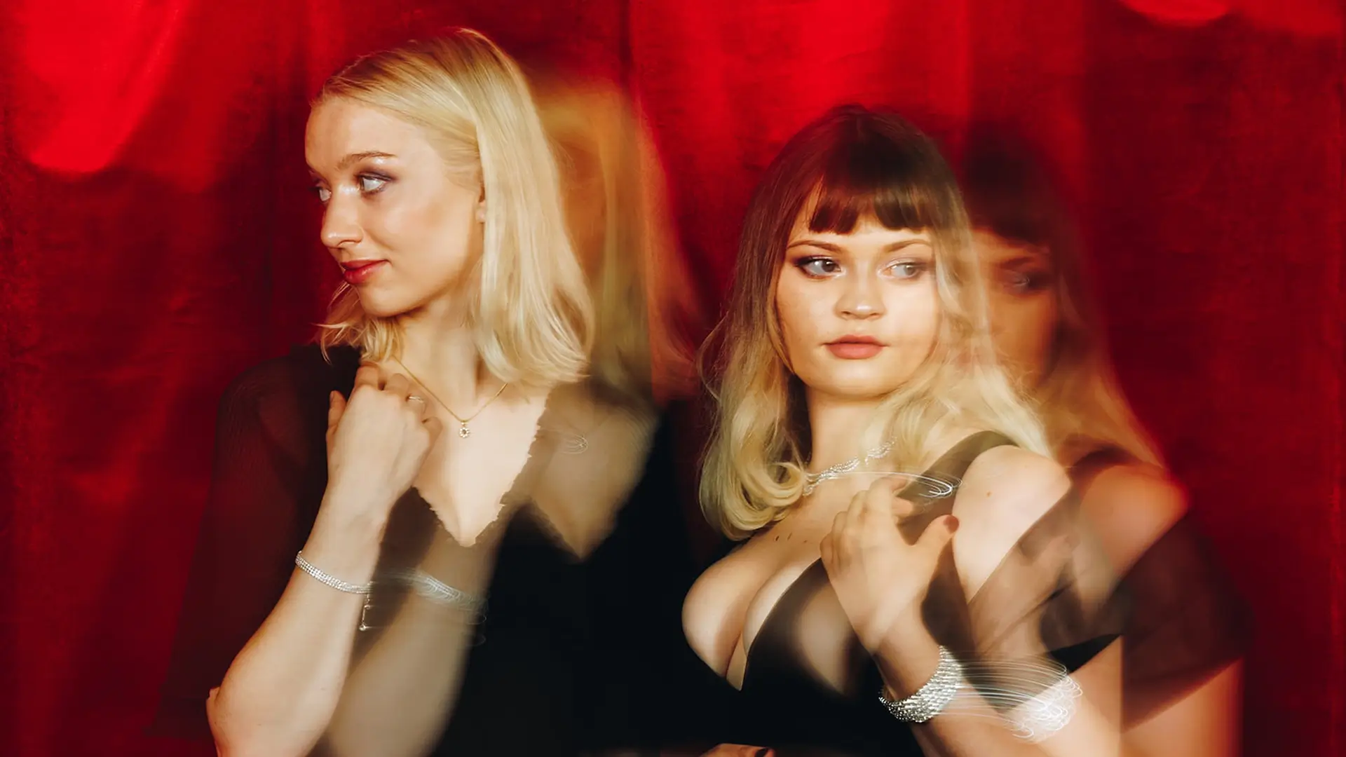 Twee zangeressen staan naast elkaar voor een rode achtergrond, stijlvol gekleed in zwart, met een dromerige en theatrale uitstraling door lichte bewegingsonscherpte.