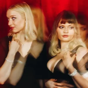 Twee zangeressen staan naast elkaar voor een rode achtergrond, stijlvol gekleed in zwart, met een dromerige en theatrale uitstraling door lichte bewegingsonscherpte.