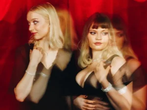 Twee zangeressen staan naast elkaar voor een rode achtergrond, stijlvol gekleed in zwart, met een dromerige en theatrale uitstraling door lichte bewegingsonscherpte.