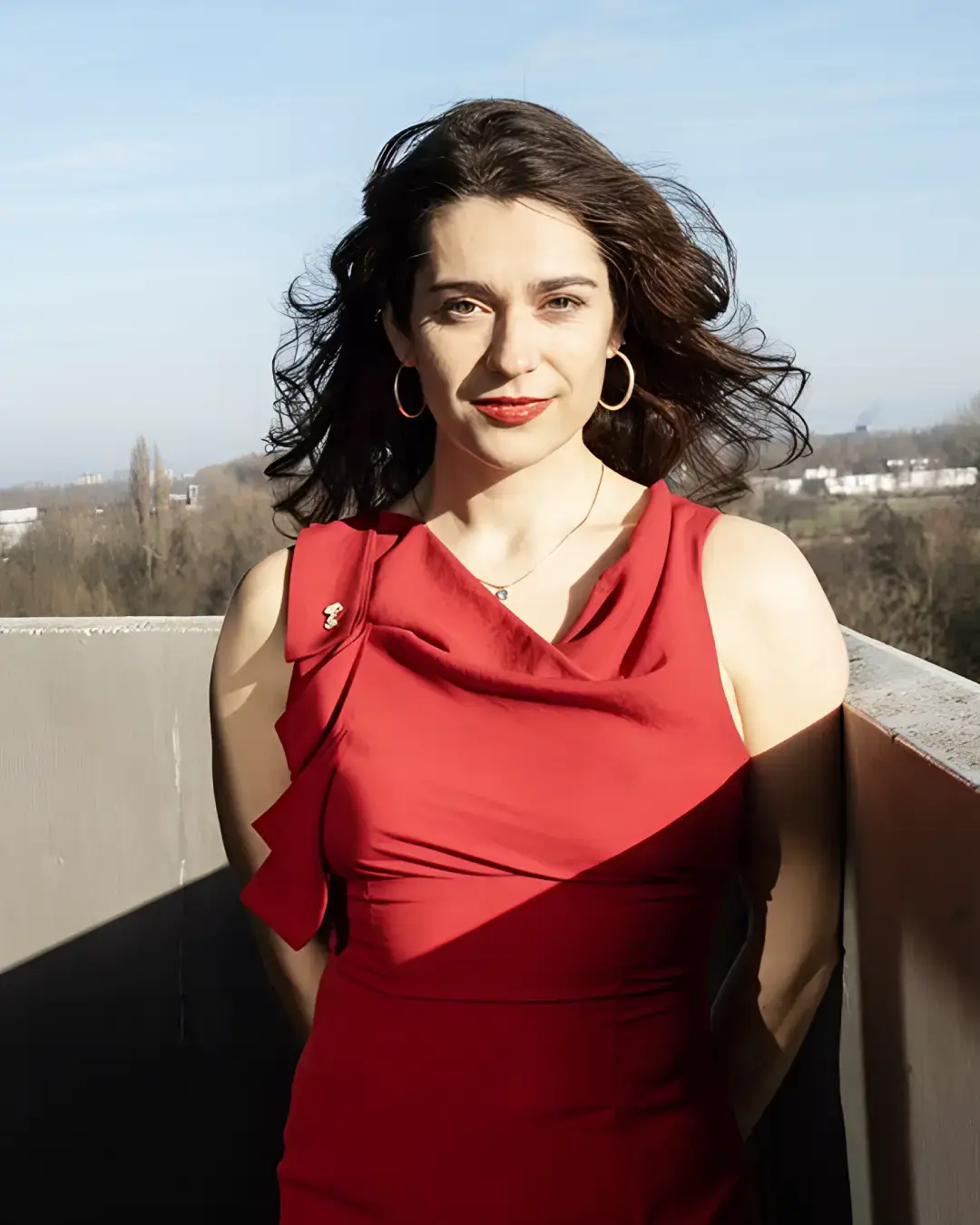 Vrouw met donker haar in een rode jurk staat buiten op een balkon, met wind door haar haar en een stadslandschap op de achtergrond.