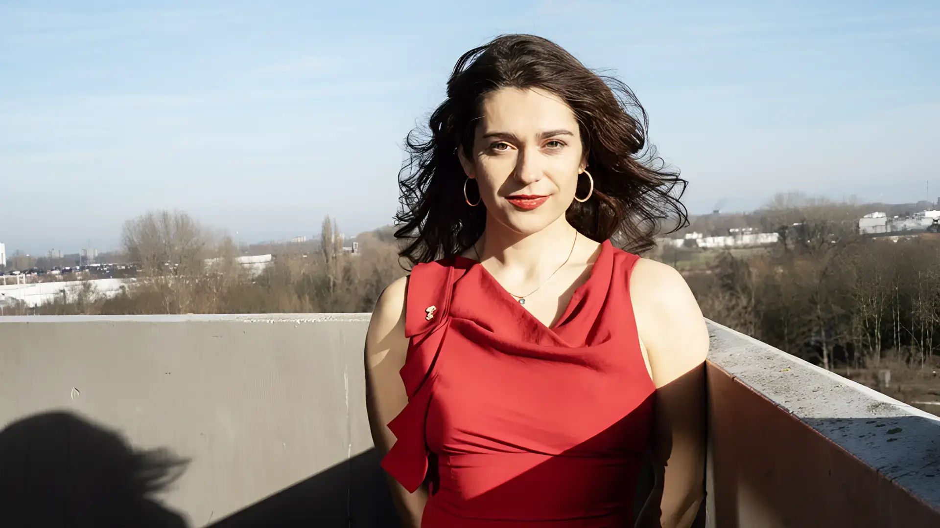 Vrouw met donker haar in een rode jurk staat buiten op een balkon, met wind door haar haar en een stadslandschap op de achtergrond.