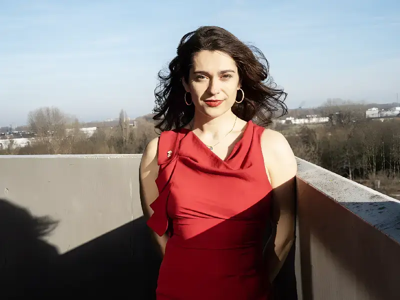 Vrouw met donker haar in een rode jurk staat buiten op een balkon, met wind door haar haar en een stadslandschap op de achtergrond.