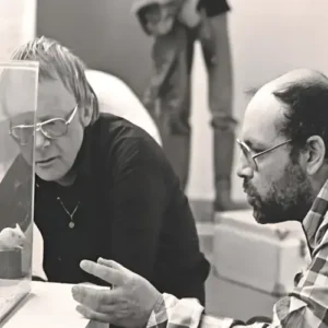 Zwart-wit foto van twee mannen die geconcentreerd kijken naar een glazen vitrine met een kleine installatie, terwijl ze met elkaar in gesprek zijn.