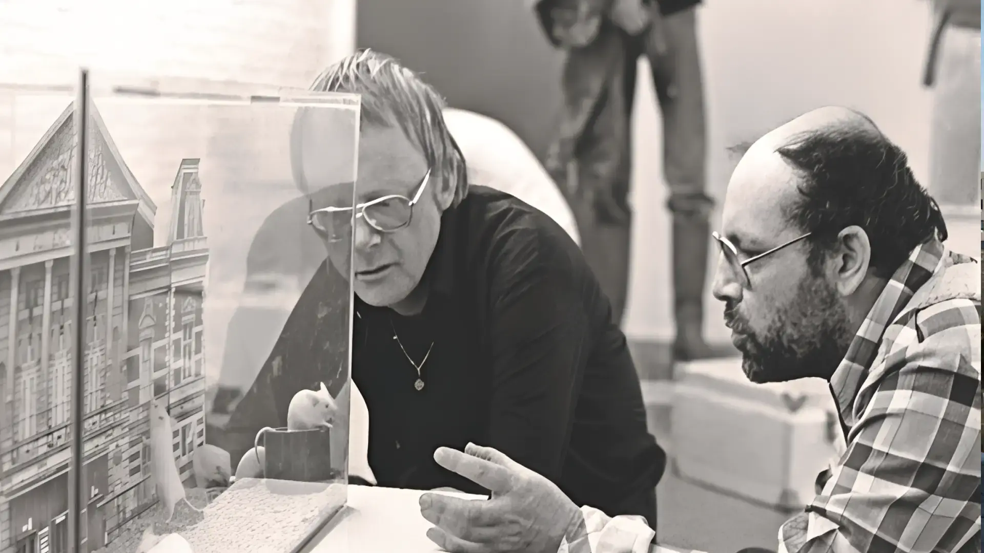 Zwart-wit foto van Misha Mengelberg & Louis Andriessen: Goede Maten die geconcentreerd kijken naar een glazen vitrine met een kleine installatie, terwijl ze met elkaar in gesprek zijn.