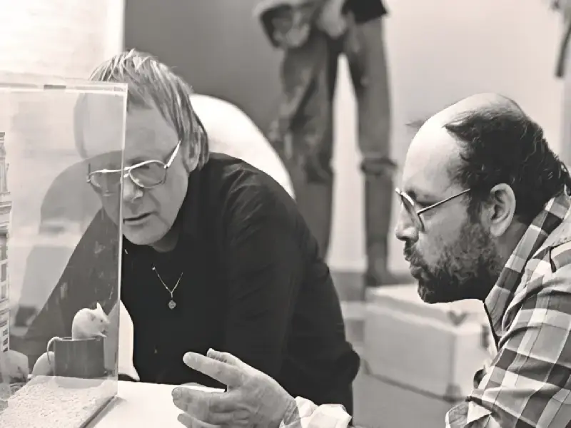 Zwart-wit foto van Misha Mengelberg & Louis Andriessen: Goede Maten die geconcentreerd kijken naar een glazen vitrine met een kleine installatie, terwijl ze met elkaar in gesprek zijn.