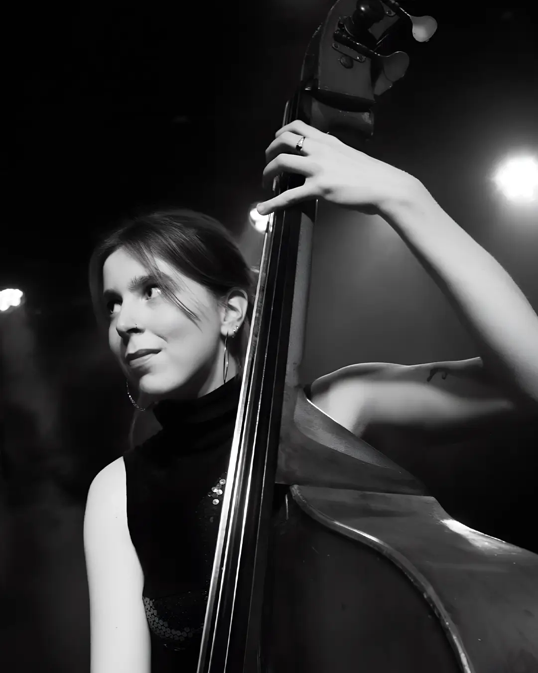 Zwart-wit portret van Veerle van der Wal - contrabassist die met een zachte glimlach opzij kijkt, terwijl ze haar instrument vasthoudt; haar gezicht is half verlicht tegen een donkere achtergrond.