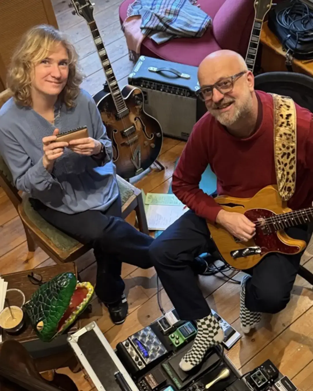 Twee muzikanten zitten in een gezellige studio: een vrouw met een mondharmonica en een man met een elektrische gitaar, omringd door instrumenten en apparatuur.