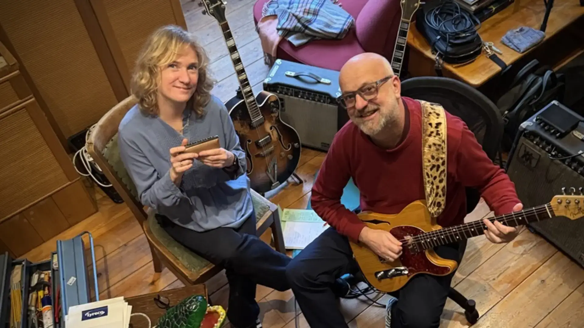 Twee muzikanten zitten in een gezellige studio: een vrouw met een mondharmonica en een man met een elektrische gitaar, omringd door instrumenten en apparatuur.
