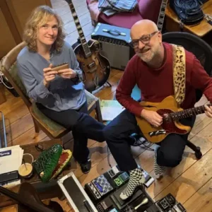 Twee muzikanten zitten in een gezellige studio: een vrouw met een mondharmonica en een man met een elektrische gitaar, omringd door instrumenten en apparatuur.