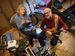 Twee muzikanten zitten in een gezellige studio: een vrouw met een mondharmonica en een man met een elektrische gitaar, omringd door instrumenten en apparatuur.