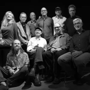 Zwart-wit groepsportret van IPC Orchestra een groot jazzensemble met mannen en één vrouw, zittend en staand op een podium tegen een donkere achtergrond.