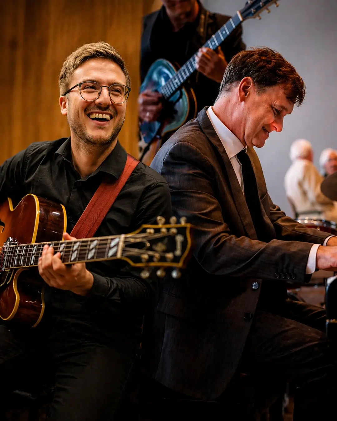 Een gitarist met bril speelt en lacht terwijl een pianist in pak geconcentreerd piano speelt tijdens een jazzoptreden.