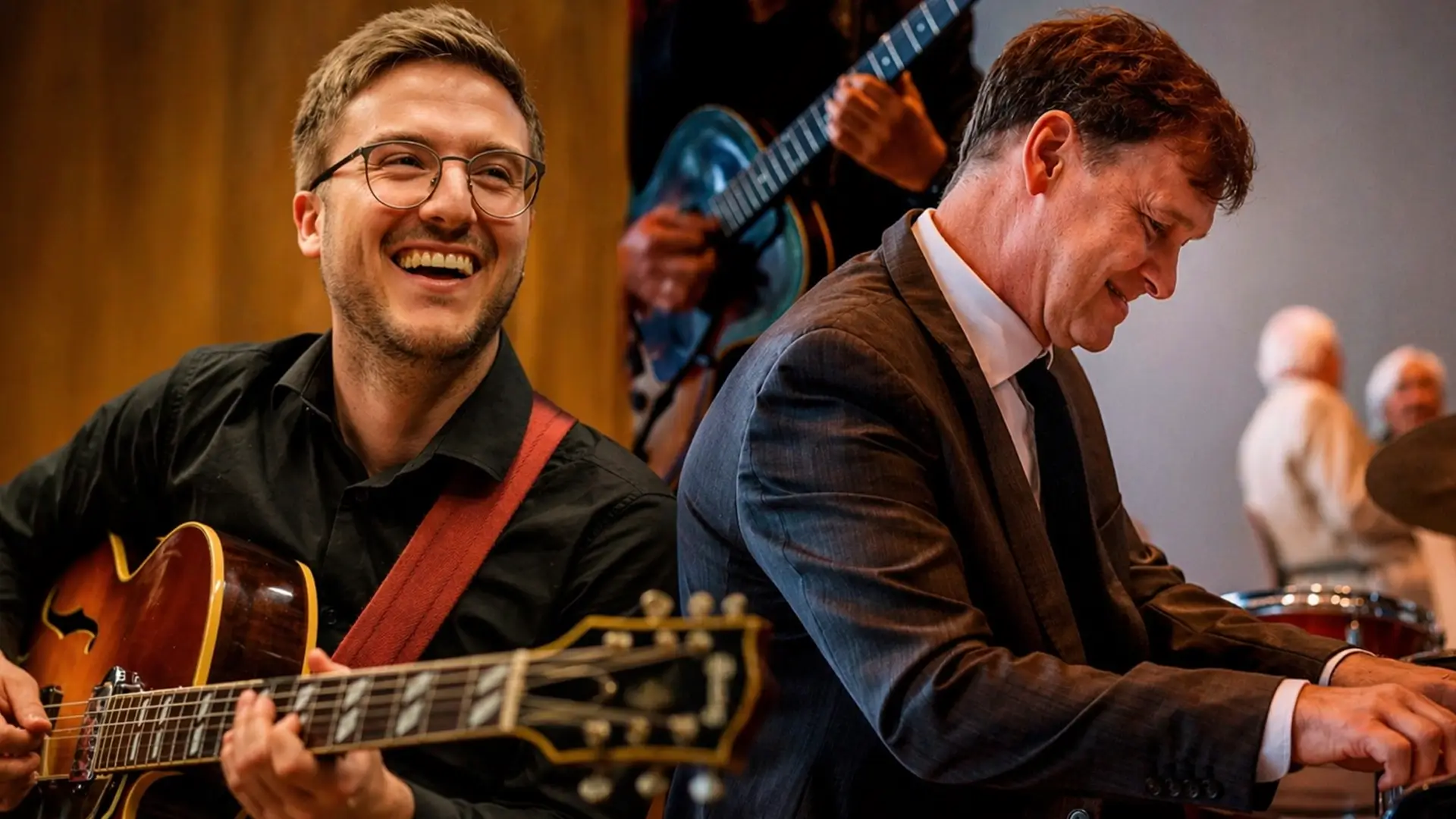Een gitarist met bril speelt en lacht terwijl een pianist in pak geconcentreerd piano speelt tijdens een jazzoptreden.