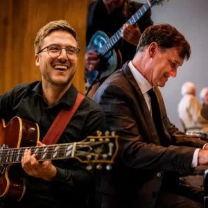 Een gitarist met bril speelt en lacht terwijl een pianist in pak geconcentreerd piano speelt tijdens een jazzoptreden.